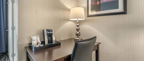 A hotel room with a desk, a lamp, a framed picture on the wall, and a coffee maker.