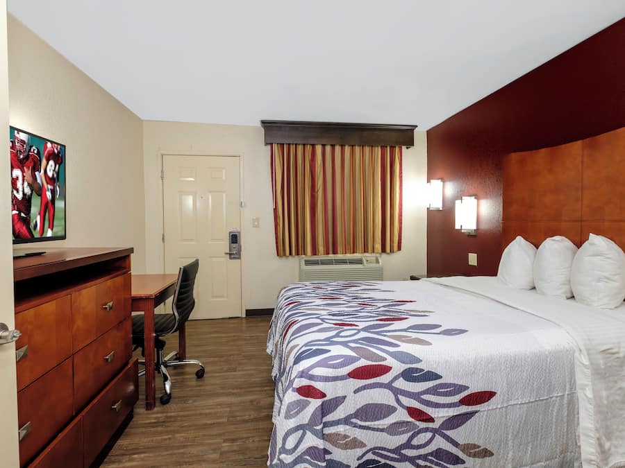 A hotel room with a bed, a desk, a chair, a dresser, a door, and a window with curtains.