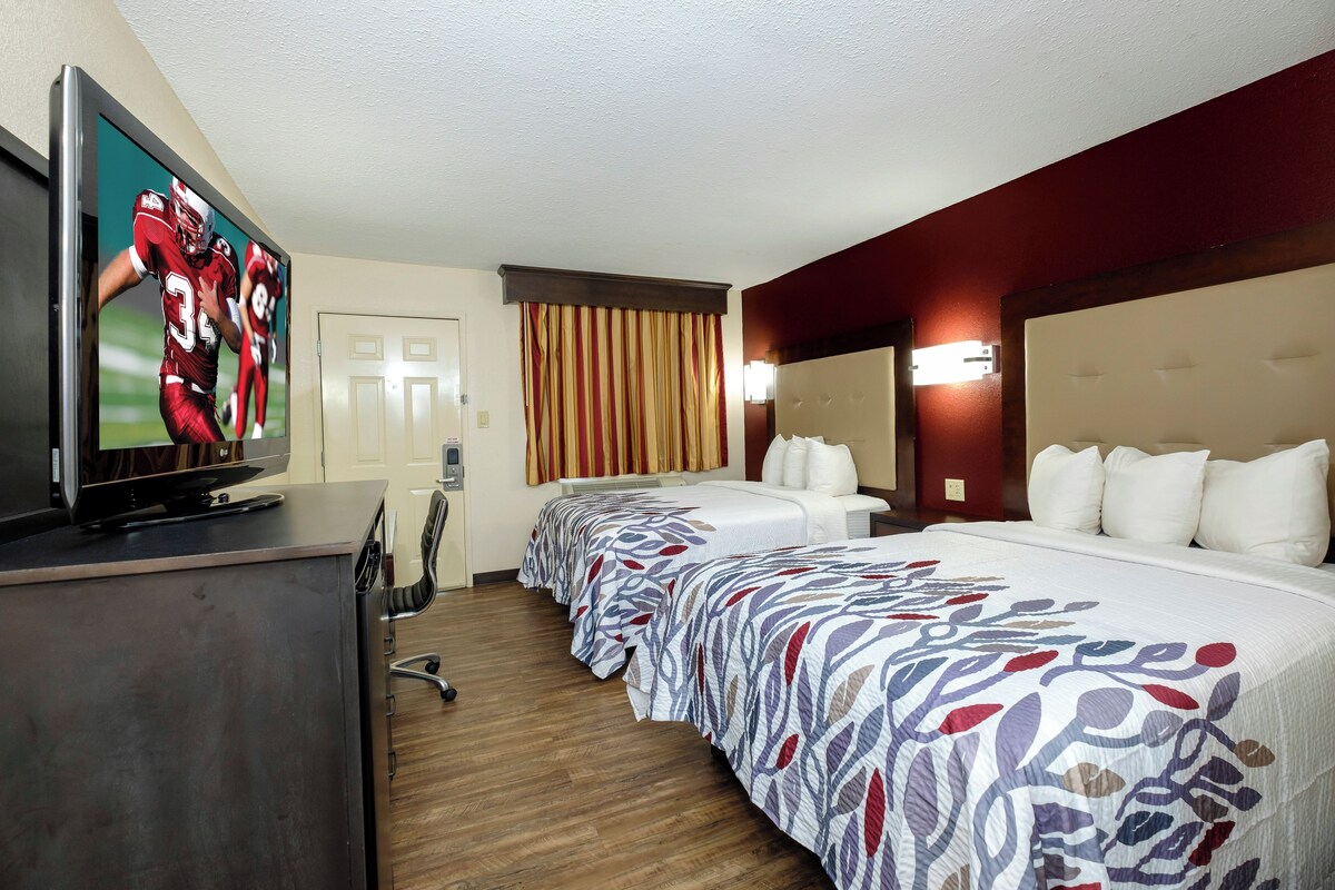 A hotel room with a television, a desk, a chair, a bed with a patterned bedspread, and a window with curtains.
