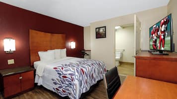 A hotel room with a bed, bedside table, lamp, TV, and a bathroom visible through an open door.