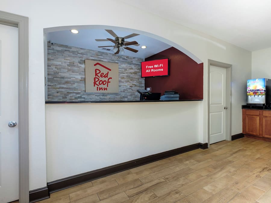 A reception area with a Red Roof Inn sign, a ceiling fan, a television displaying 'Free Wi-Fi All Rooms', and a coffee machine.