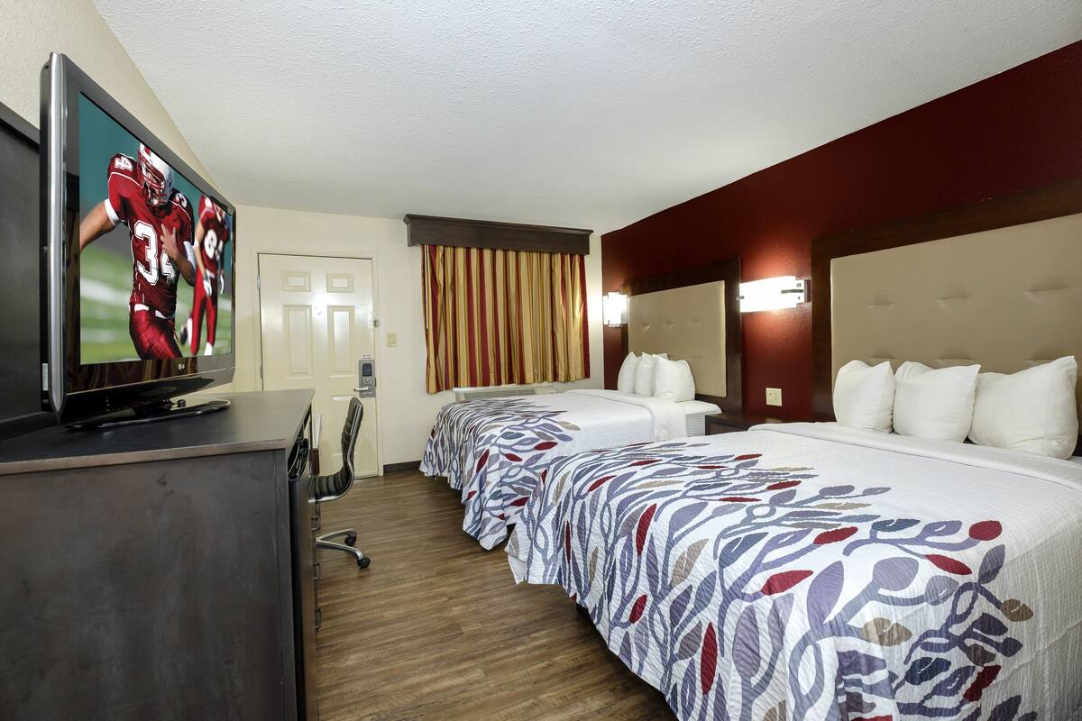 Hotellihuone, jossa on työpöydällä televisio, kuviollinen päiväpeite ja päätyosa varustettu sängyllä sekä seinään kiinnitetty valaisin.