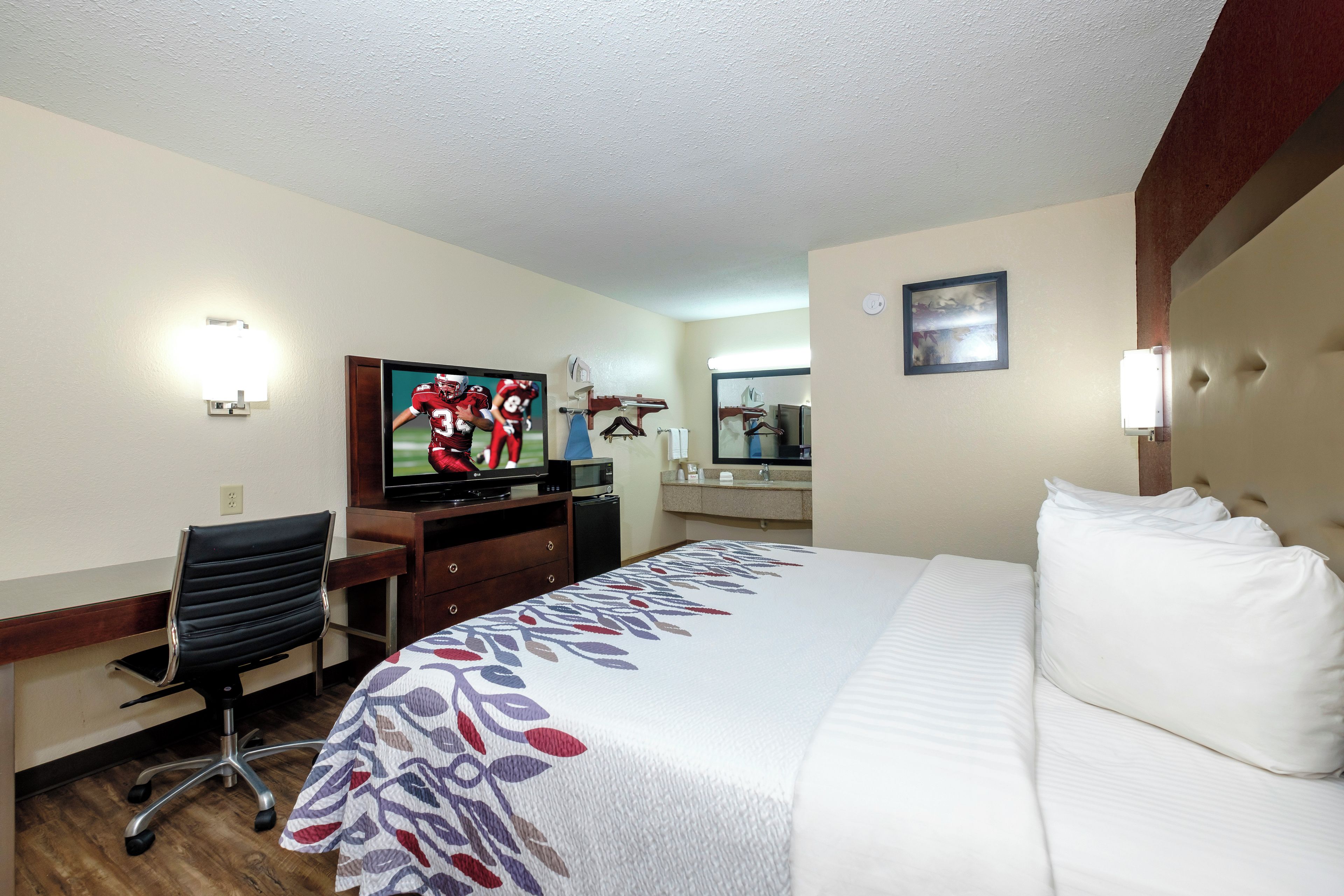 A hotel room with a bed, a desk with a chair, a television, and a bathroom mirror.