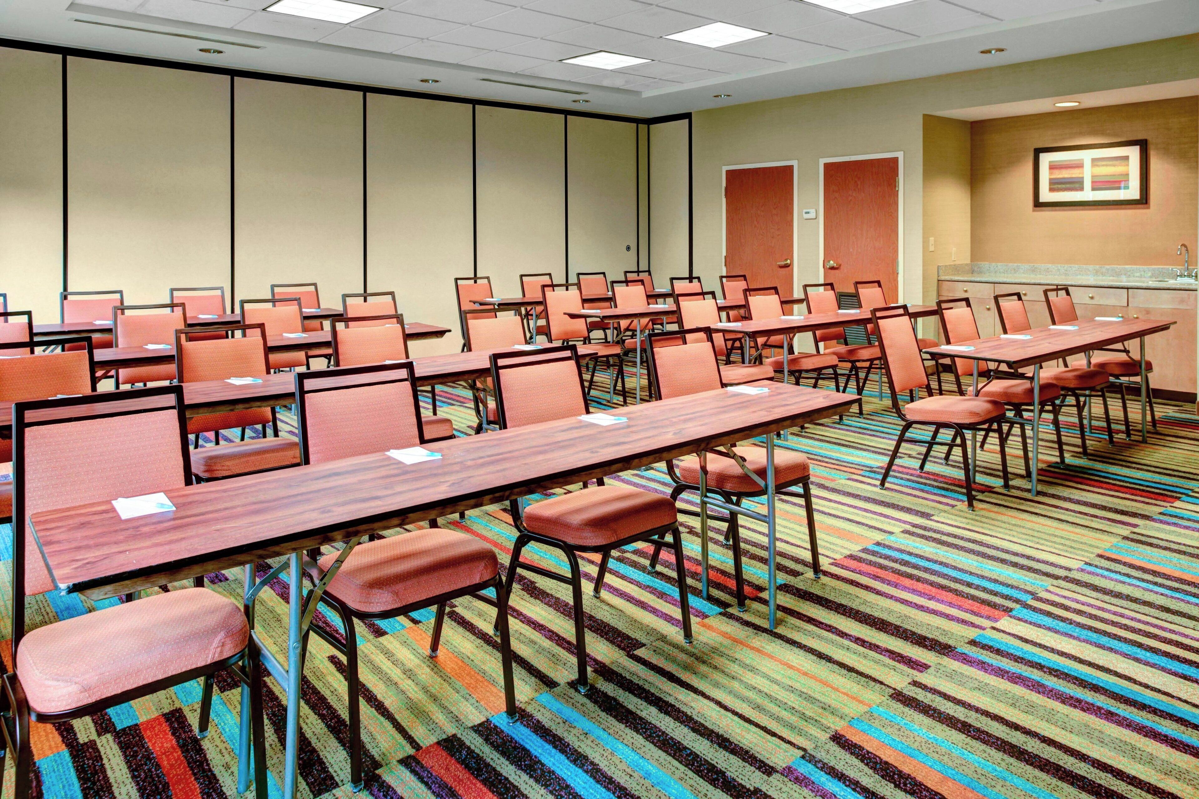A conference room with multiple tables and chairs, a carpeted floor, and a kitchenette area.