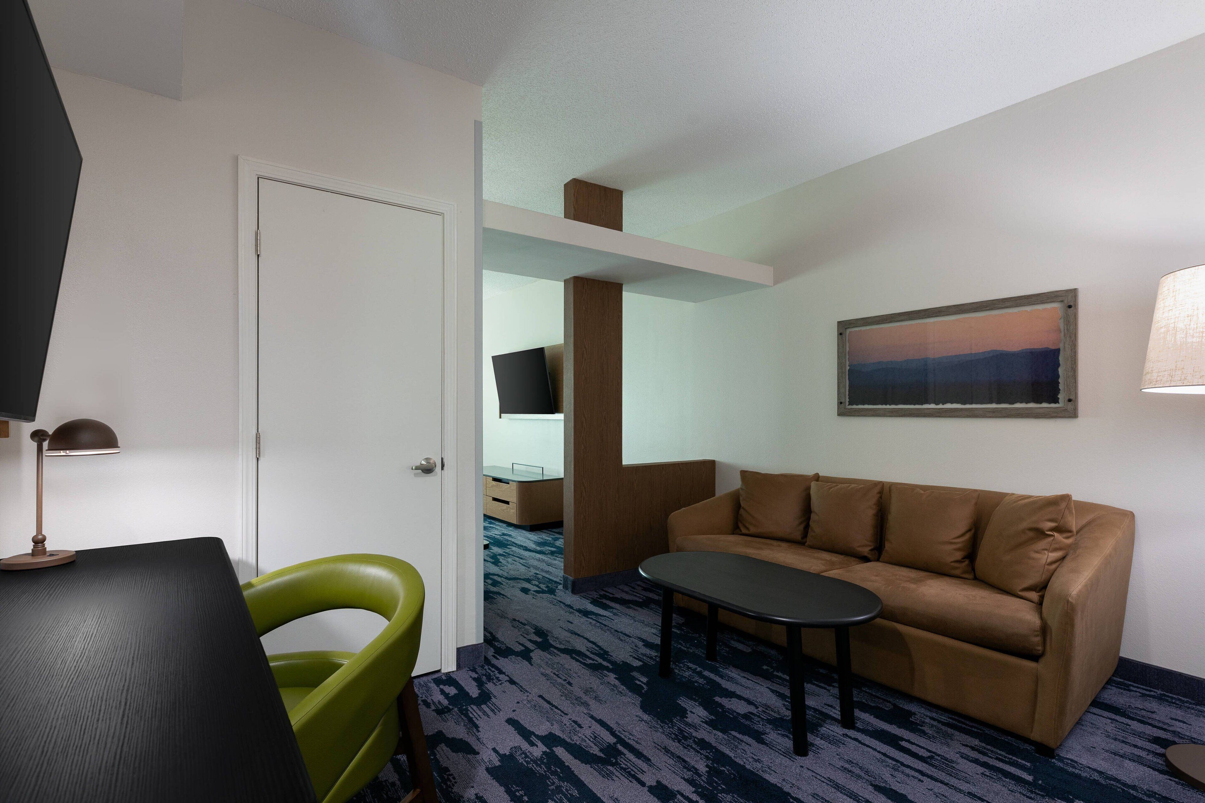 A hotel room with a brown sofa, a small round table, a green chair, a desk with a lamp, and a painting on the wall.