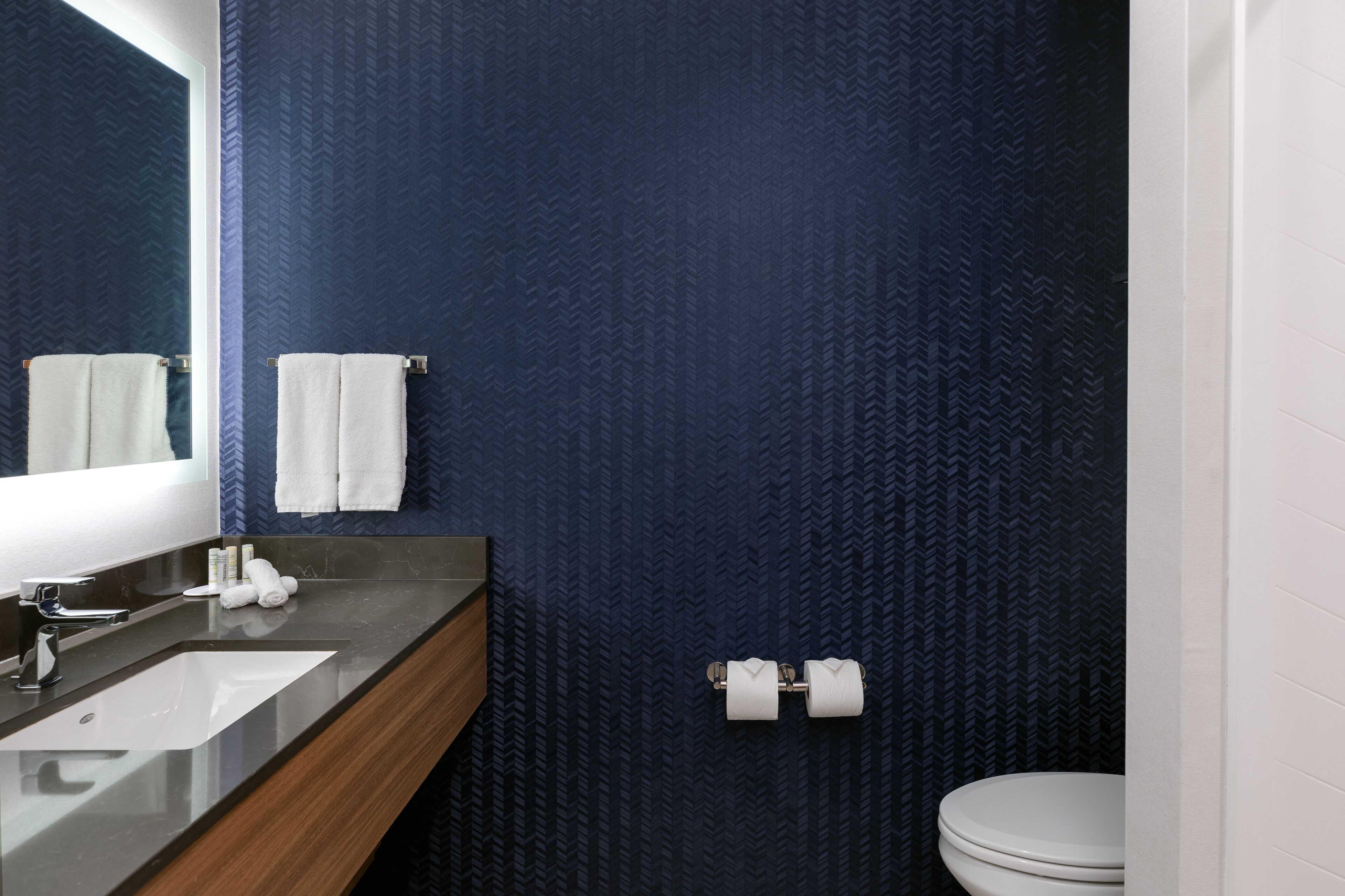 A bathroom with a dark blue textured wall, a white sink with a wooden vanity, and a toilet with two rolls of toilet paper.