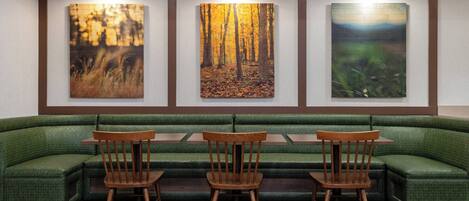 A restaurant with a green cushioned bench seating area, wooden chairs, and three framed artworks on the wall.