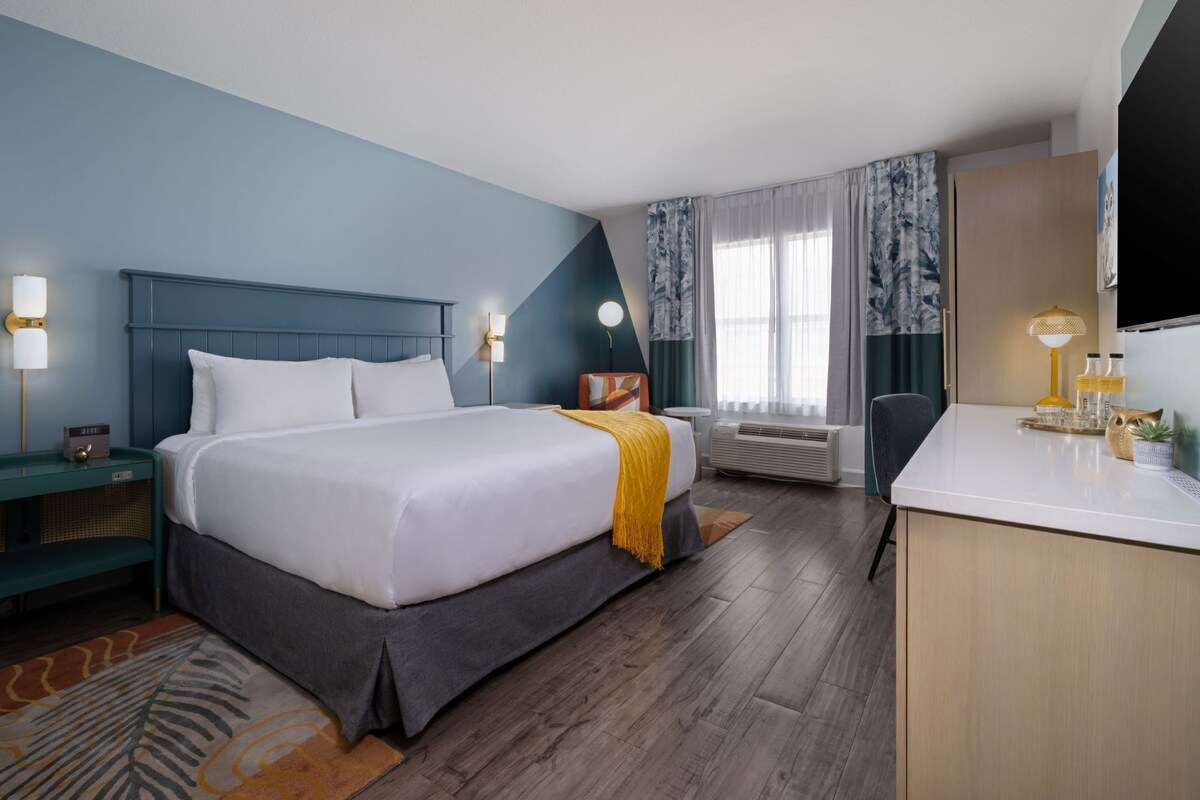 Ein Hotelzimmer mit Bett, Nachttisch, Lampe, Klimaanlage, Schreibtisch, Stuhl und Fernseher.