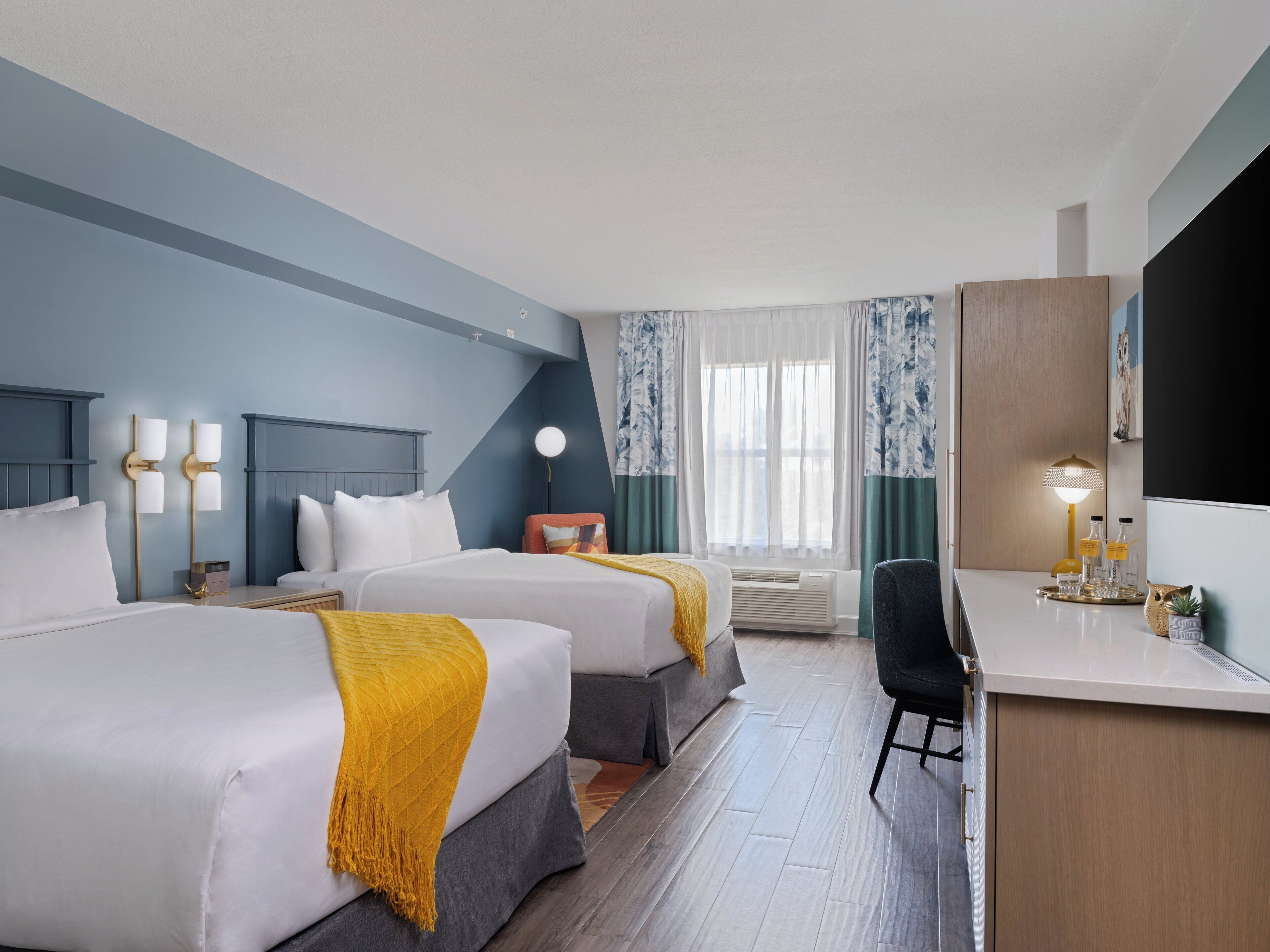 A hotel room with two beds, a desk, a chair, a lamp, and a large mirror.