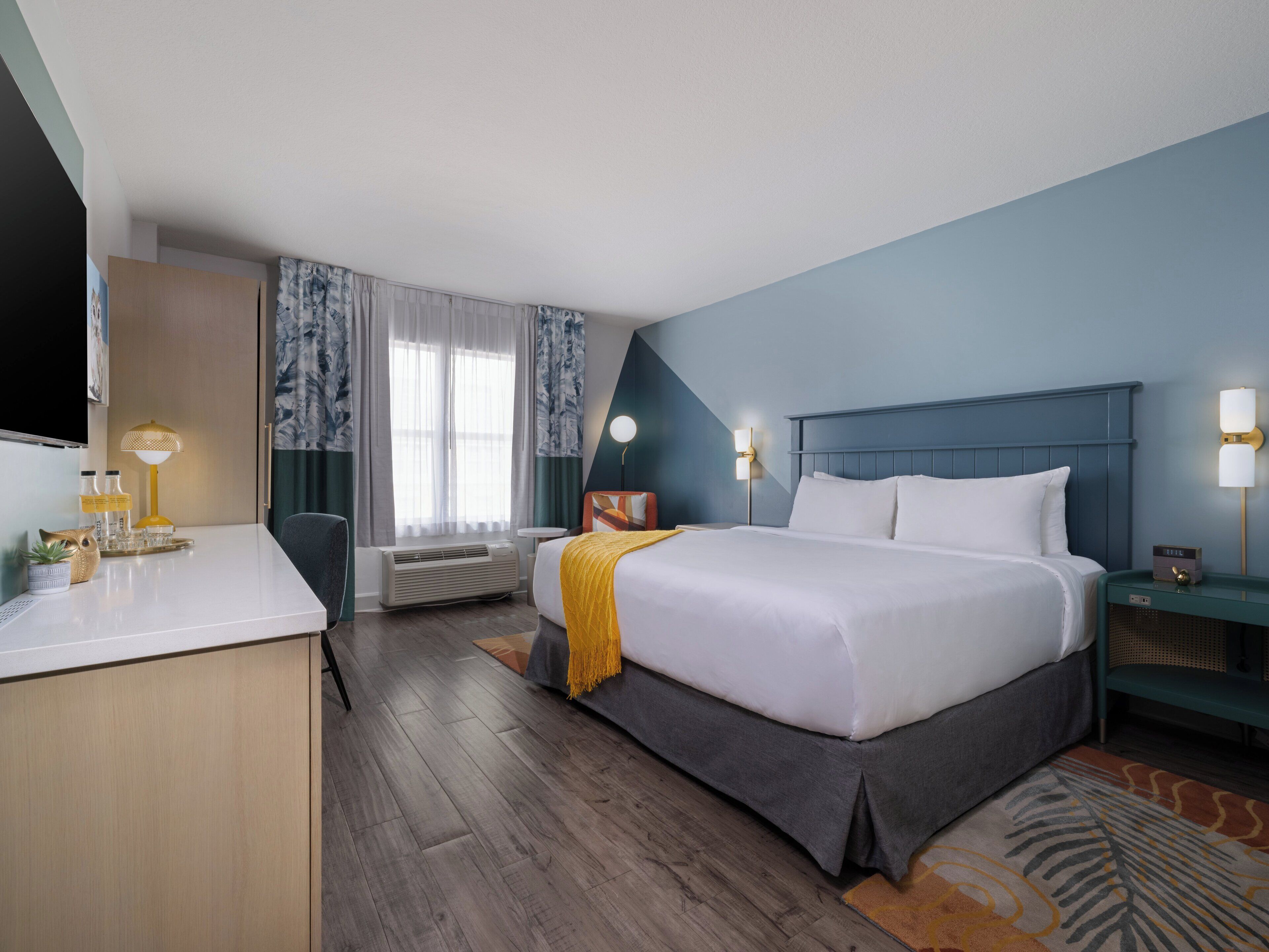 A hotel room with a bed, bedside table, lamp, air conditioning unit, and a desk with a chair.