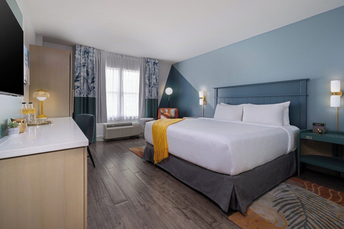 A hotel room with a bed, bedside table, lamp, air conditioning unit, and a desk with a chair.