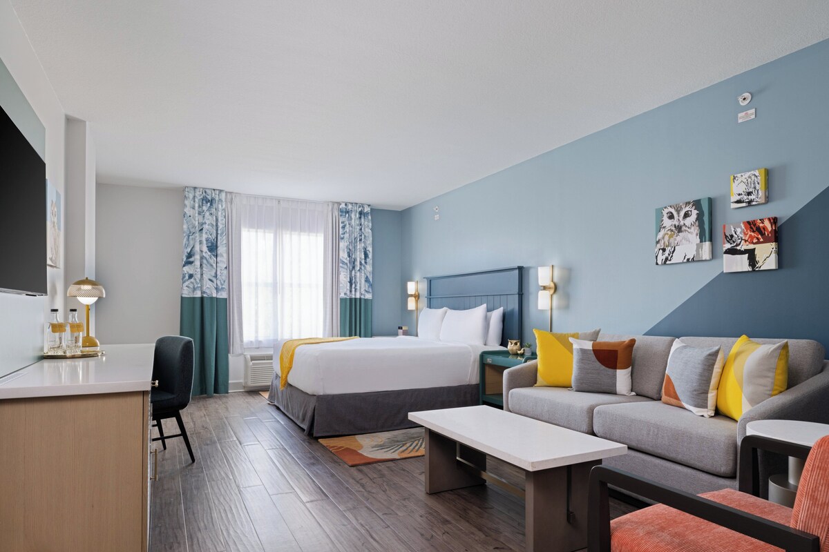 A modern hotel room with a bed, sofa, desk, and a view of the city.