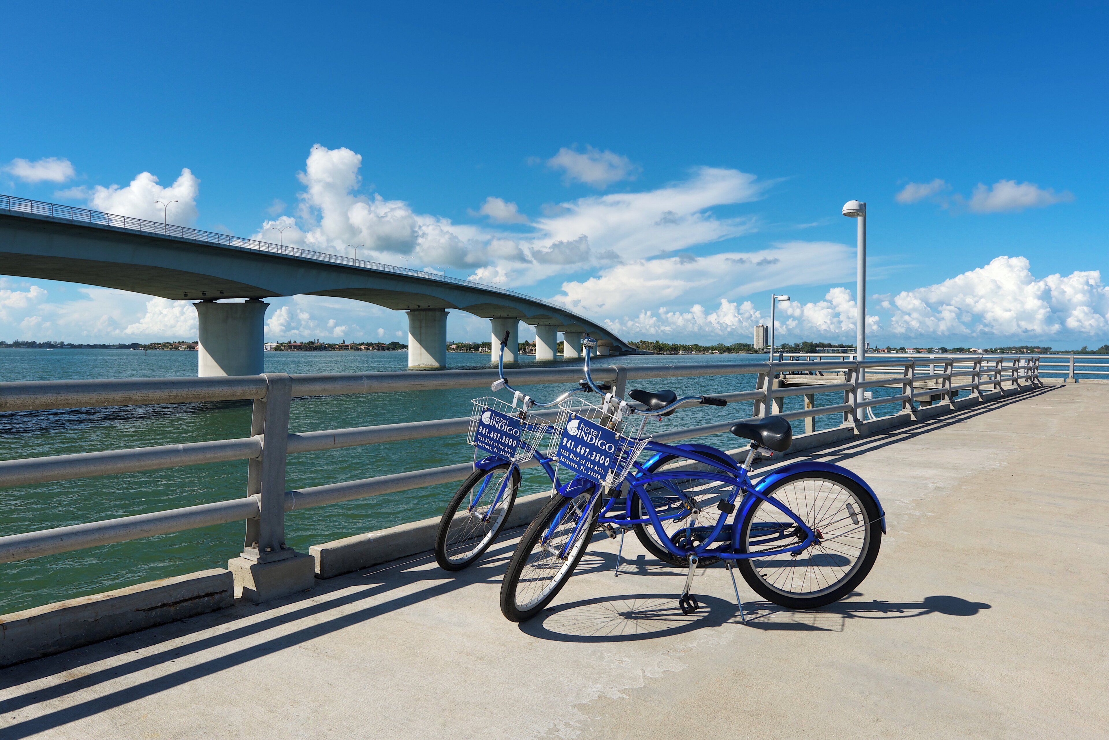 A blue bicycle with a hotel sign attached to it, parked on a concrete walkway by a waterfront.