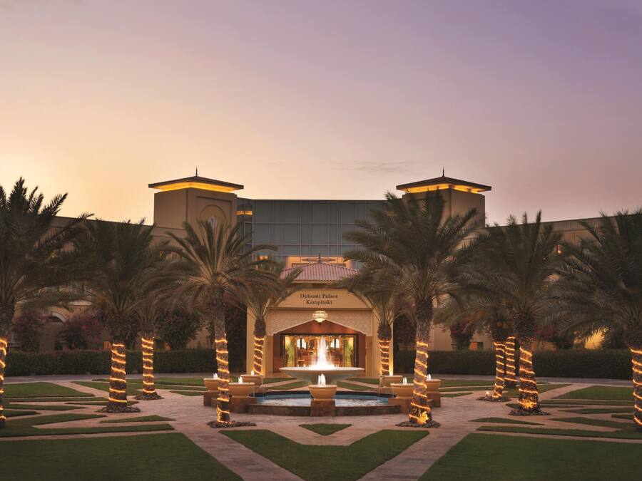 A hotel entrance with a fountain, palm trees, and a well-maintained garden.