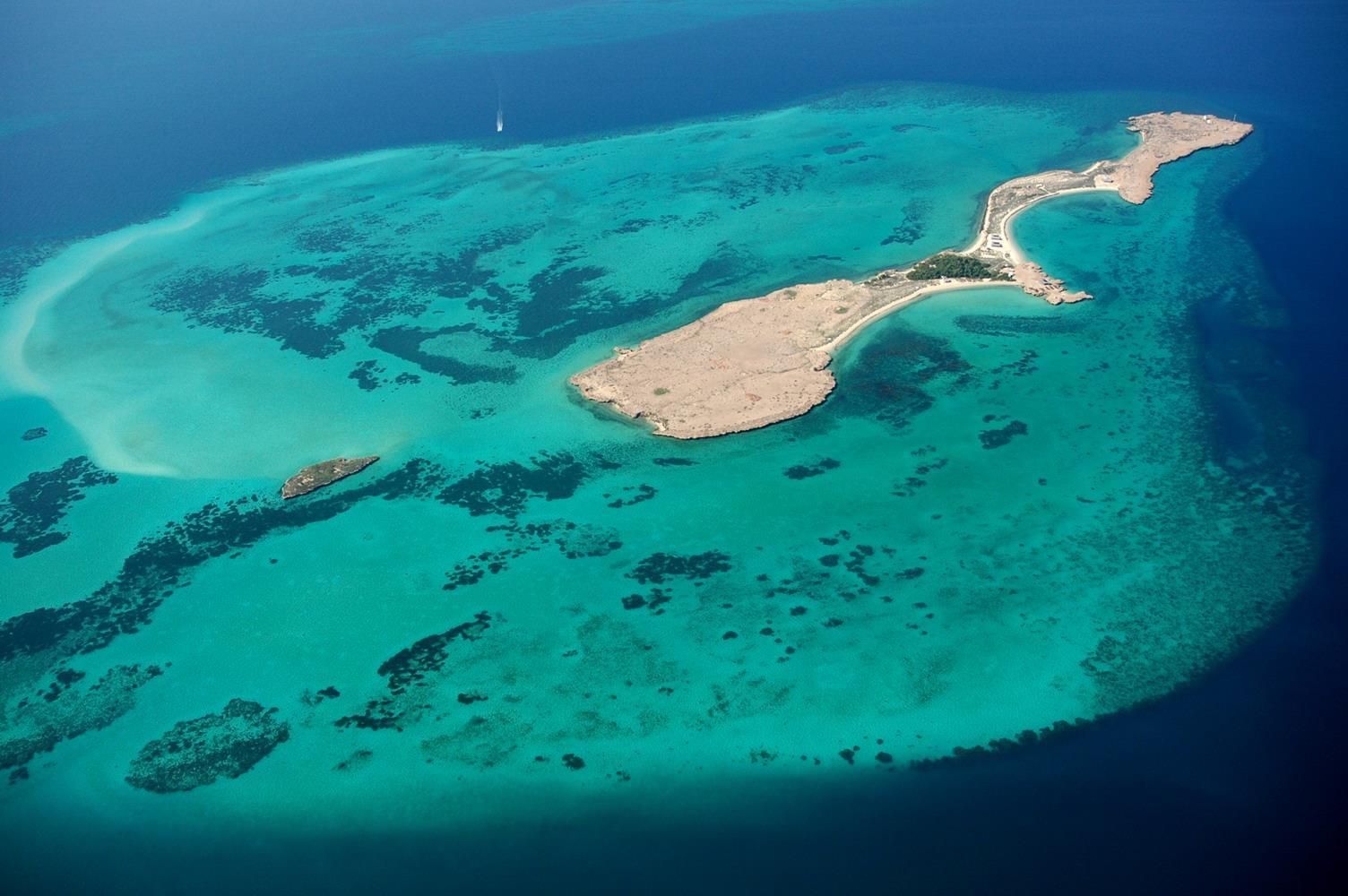 Eine Vogelperspektive auf eine Küstenregion mit klarem türkisblauem Wasser, einem Sandstrand und einer kleinen Insel, die durch einen schmalen Landstrich mit dem Festland verbunden ist.