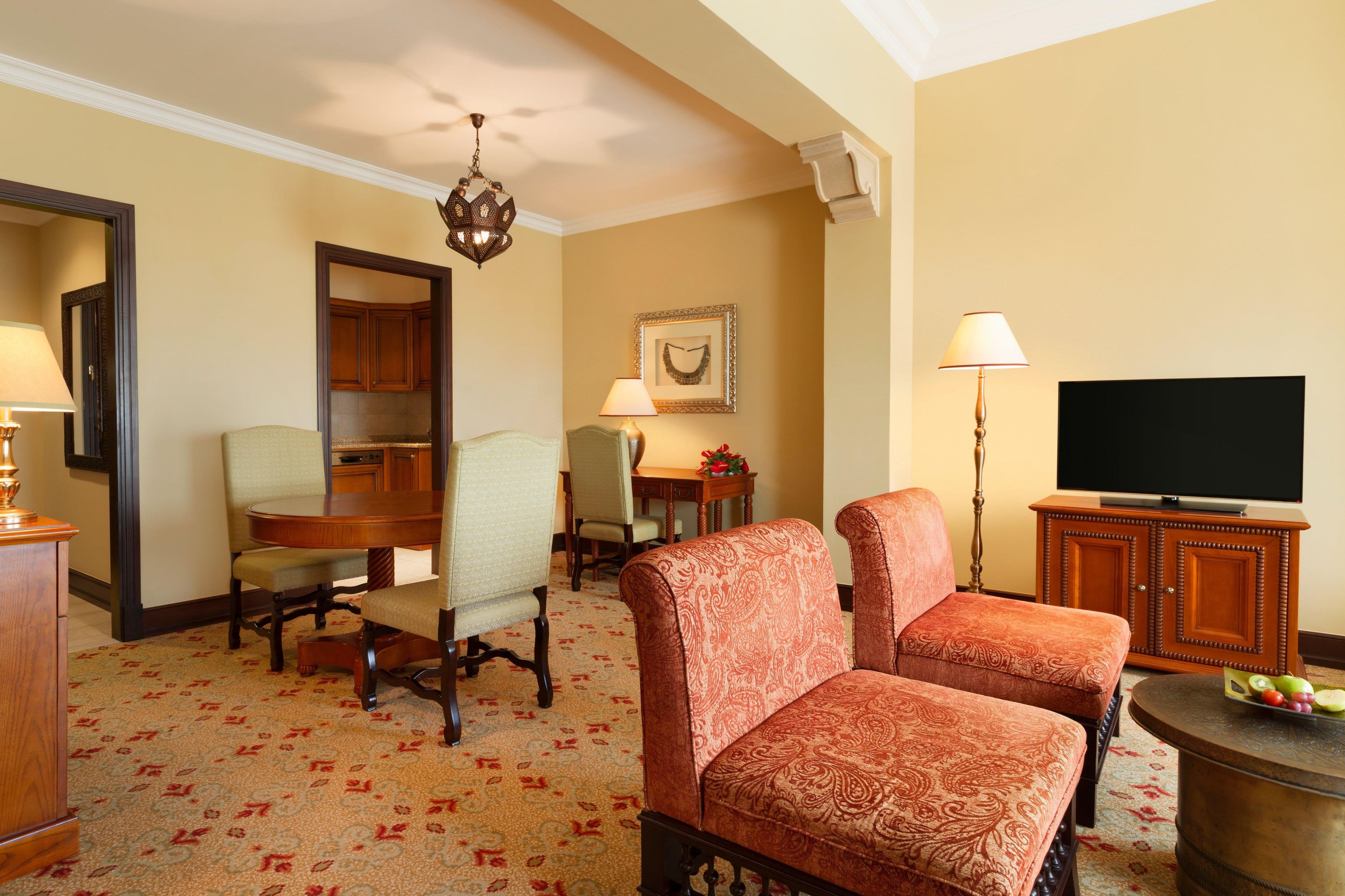 Quarto de hotel com uma mesa de jantar, duas poltronas, uma televisão e um espelho.