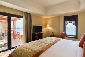 A hotel room with a bed, a TV, a lamp, a chair, and a view of the sea through a window. - Djibouti Palace Kempinski (Djibouti)