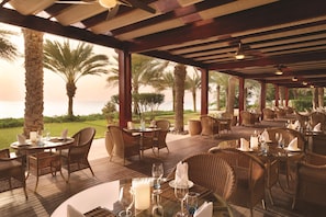 An outdoor dining area with tables and chairs, palm trees, and a view of the sea. - Djibouti Palace Kempinski (Djibouti)