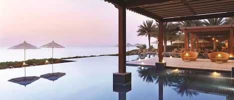 Una piscina infinita tranquila con agua cristalina que refleja el cielo, rodeada de sillones de descanso y sombrillas, y vistas al mar al atardecer.