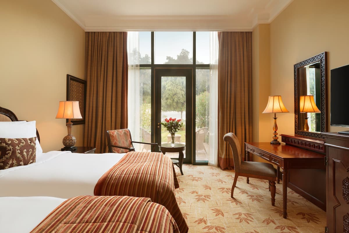 A hotel room with two beds, a desk, a chair, a TV, and a view of the outdoors through a glass door.