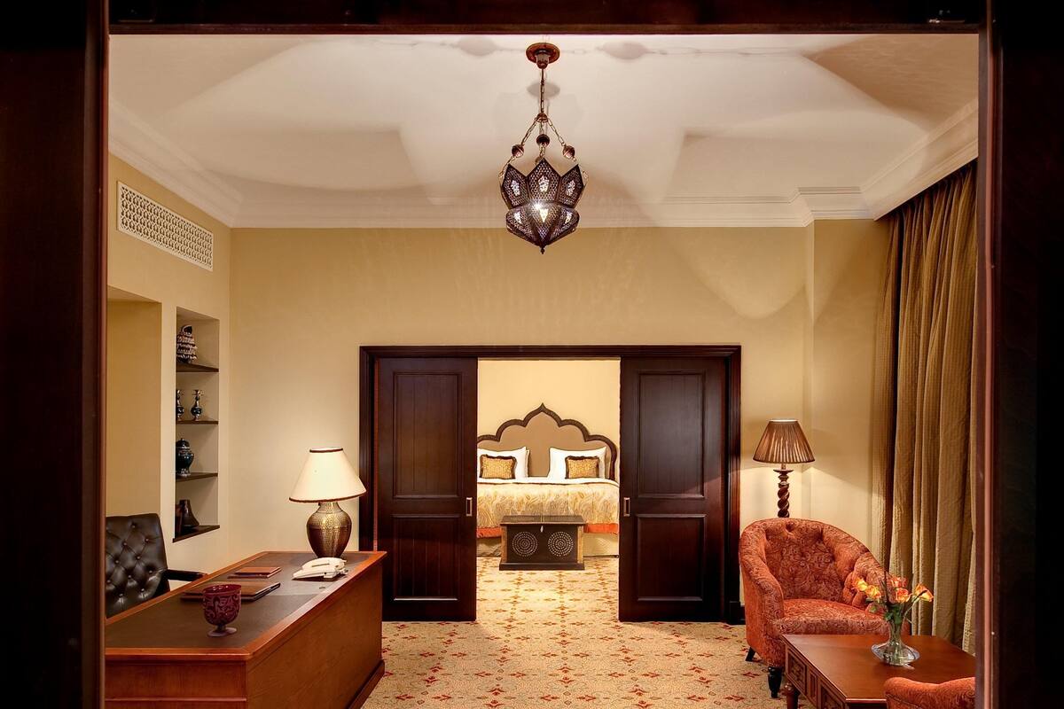 A hotel room with a wooden desk, a chair, a bed with a headboard, a nightstand, and a lamp.