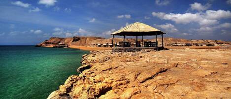 A rocky coastline with a thatched-roof structure, clear turquoise water, and distant hills.