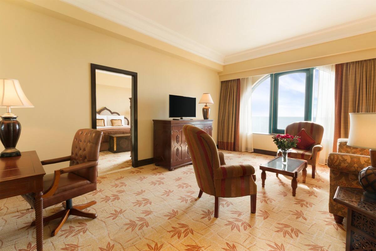 A hotel room with a large window, a television, a desk, and a seating area with a patterned carpet.