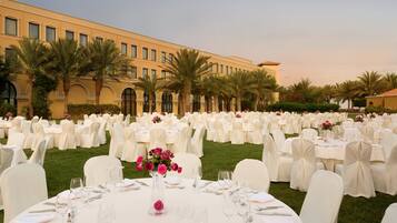 Un salón de banquetes al aire libre con filas de mesas redondas dispuestas para un evento formal, rodeado de palmeras y un edificio grande al fondo.