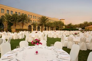 An outdoor banquet hall with rows of round tables set for a formal event, surrounded by palm trees and a large building in the background. - Djibouti Palace Kempinski (Djibouti)