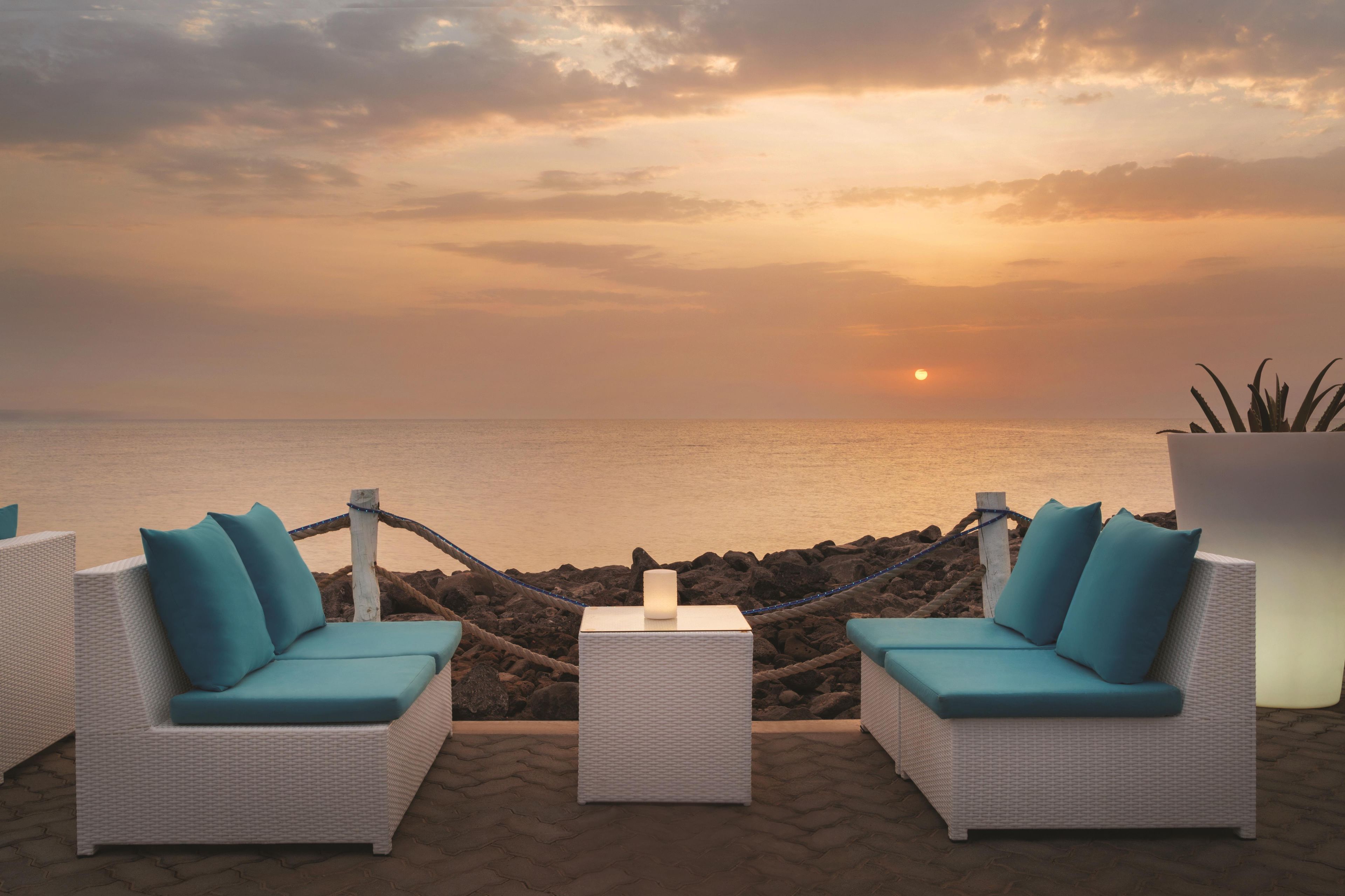 A seaside lounge with two white wicker sofas with teal cushions, a small white table, and a potted plant.