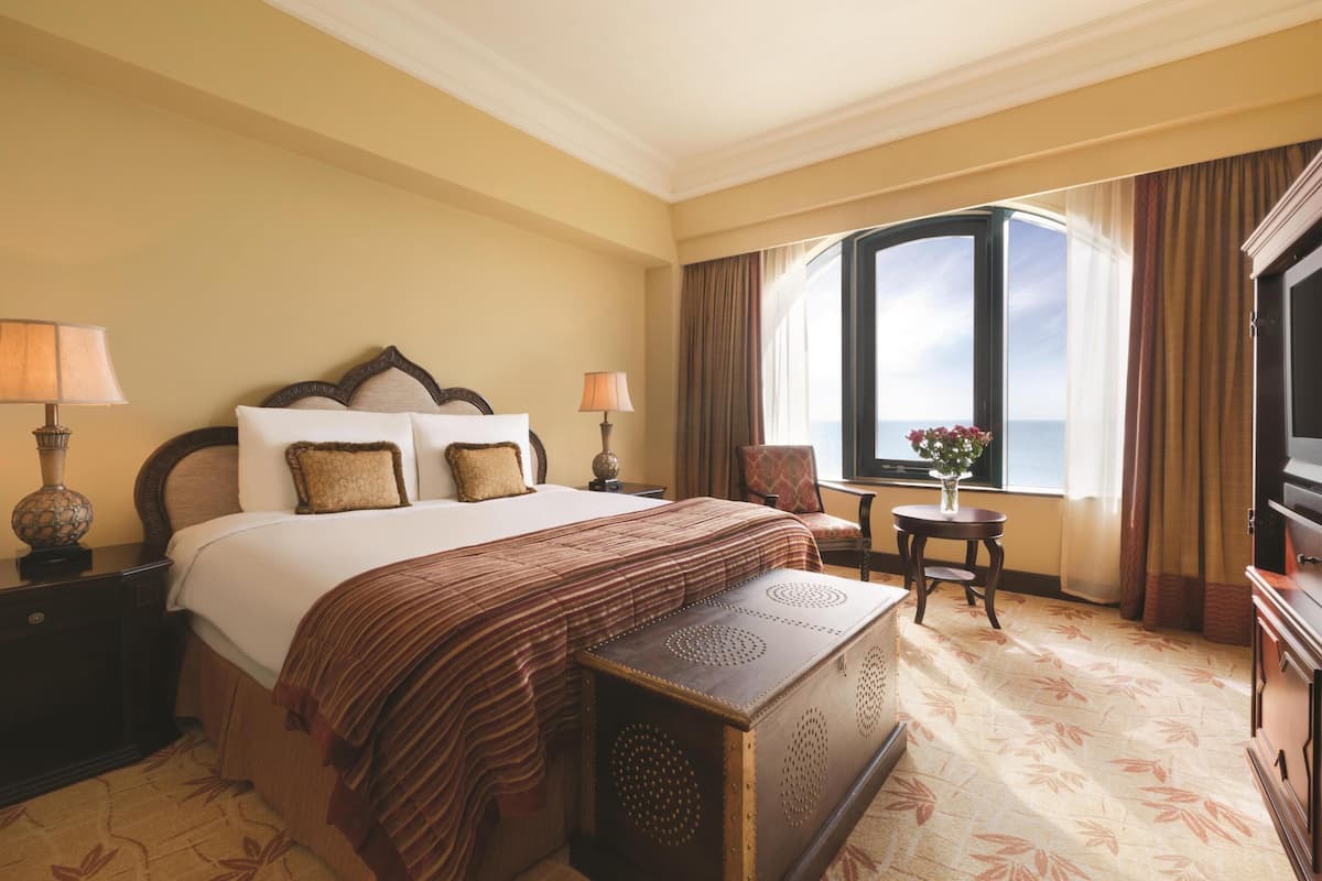 A hotel room with a large bed, two bedside tables with lamps, a small table with a vase, a television, and a window with curtains.