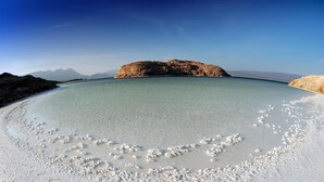 En strand med klart vatten, en sandstrand och en stor klippformation i bakgrunden.