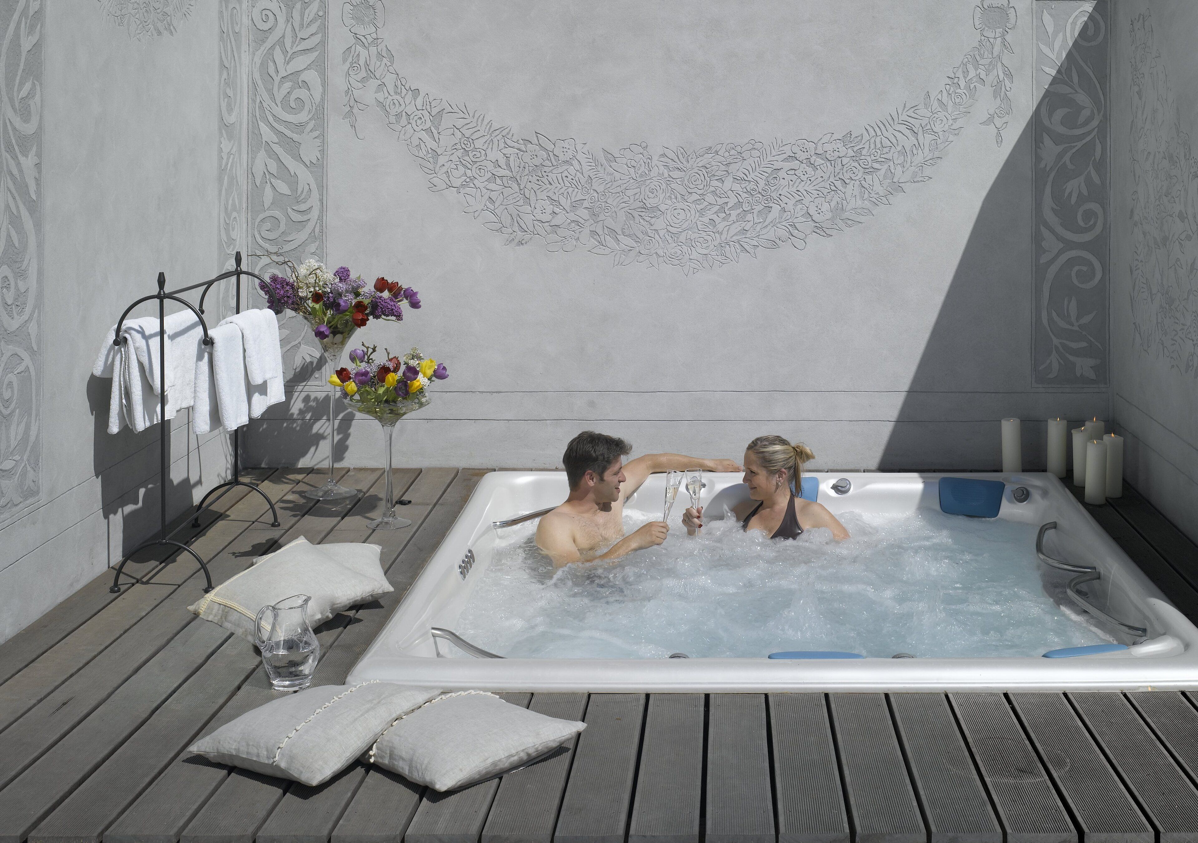 Un couple dans un bain à remous, entouré de serviettes et de fleurs, sur une terrasse en bois.