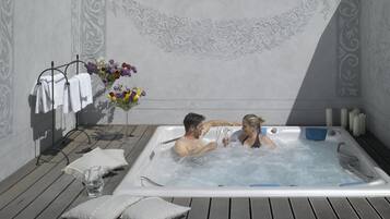 Un couple dans un bain à remous, entouré de serviettes et de fleurs, sur une terrasse en bois.