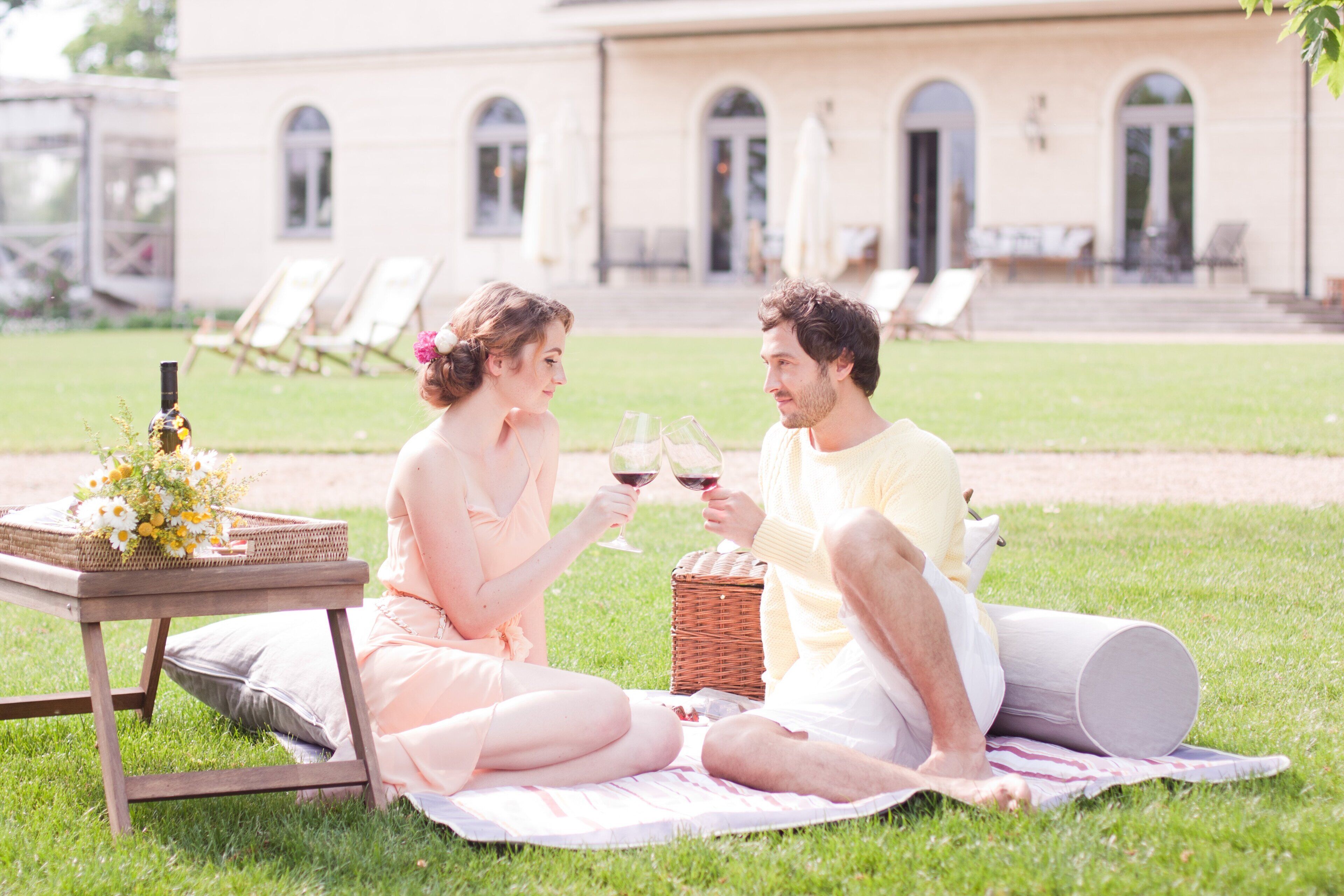A couple sitting on a blanket outdoors with a table between them, a wicker basket, and a bottle of wine.