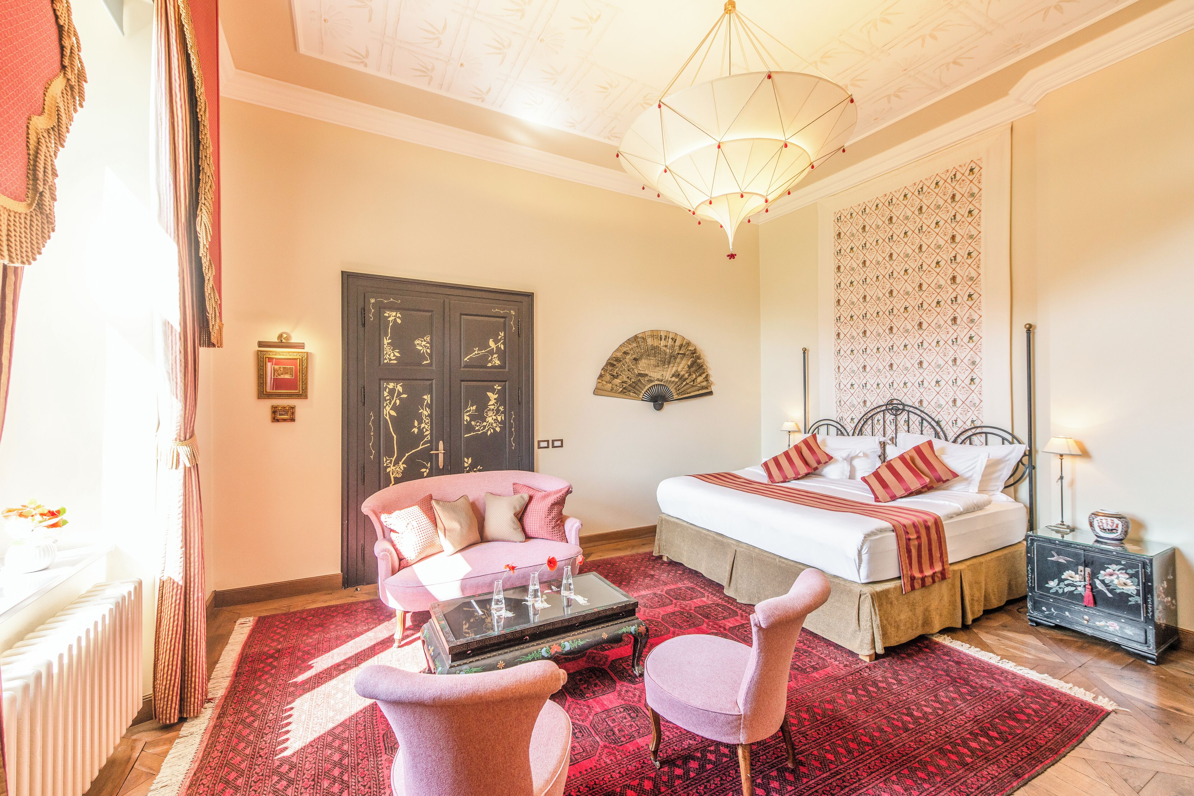 Un dormitorio con cama, una zona de estar con sofá y sillones, un abanico decorativo y una alfombra roja estampada.