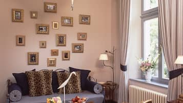 Una habitación con sofá, sillas, una mesa con flores, cuadros en la pared y una ventana con cortinas.