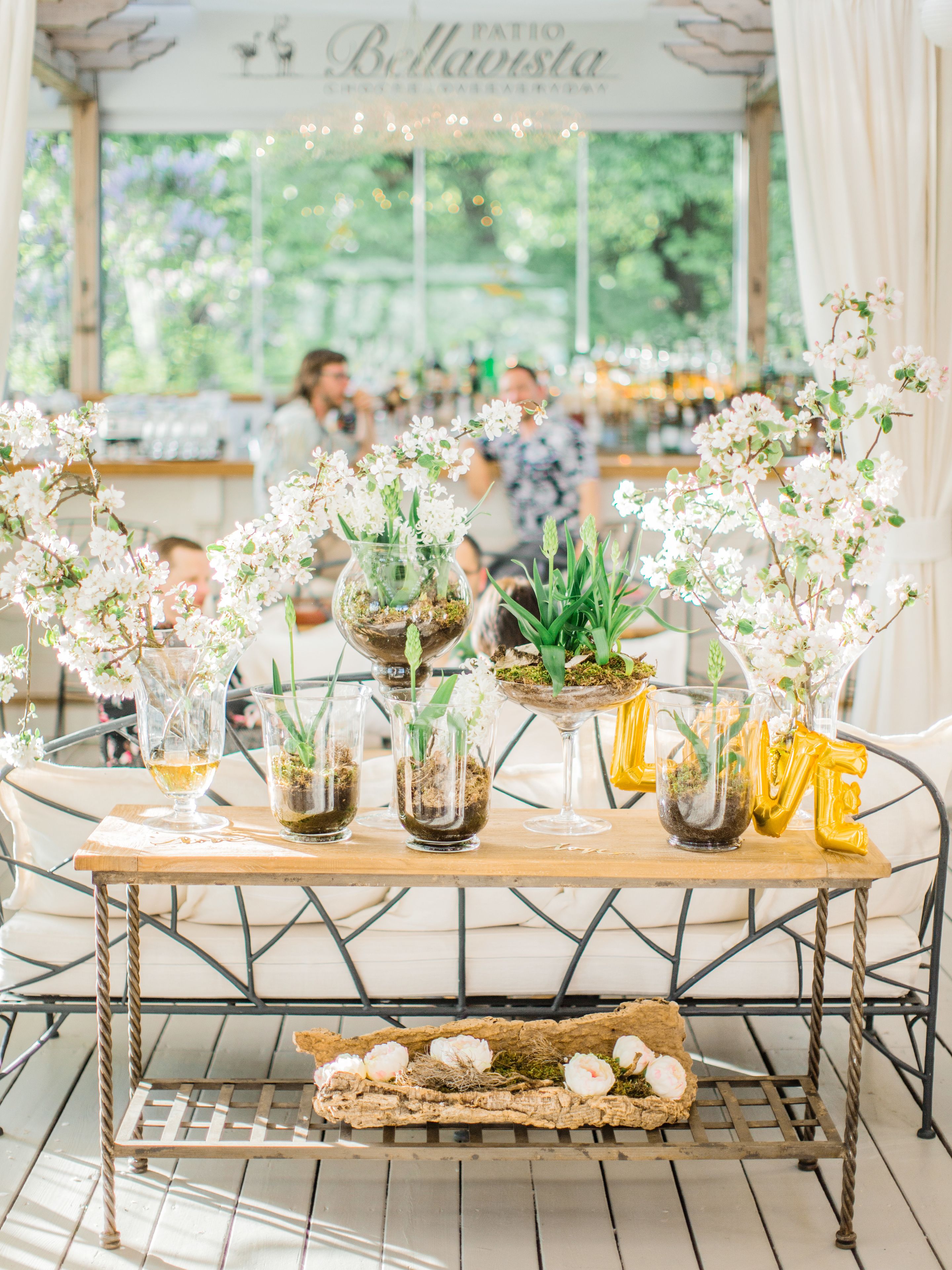 A table with glass vases containing flowers and plants, a tray with decorative items, and a sign above reading "Patio Bellavista".