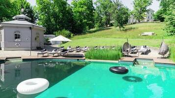 A swimming pool with lounge chairs and a small building, surrounded by greenery and a hill.