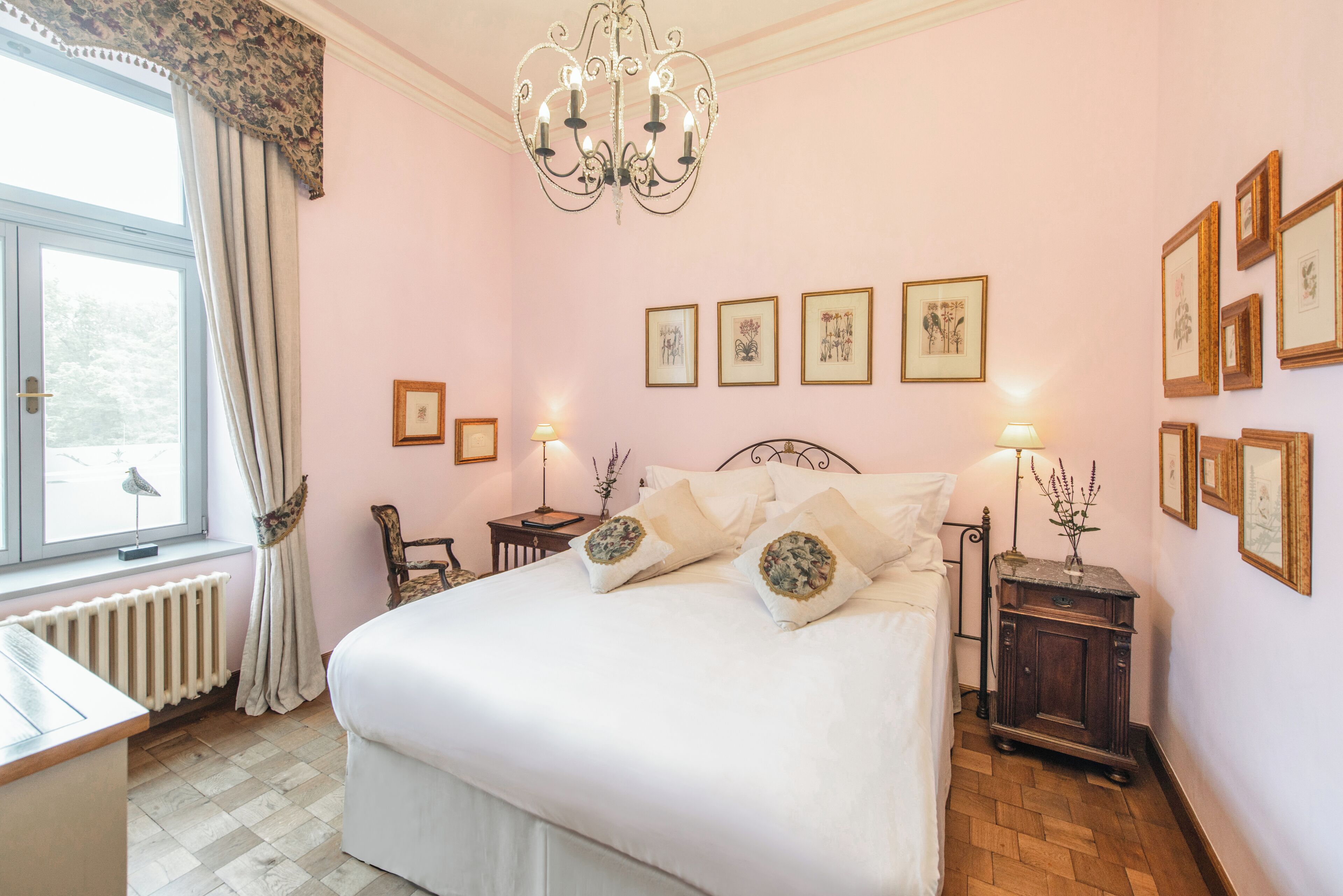 A bedroom with a large bed, a chandelier, framed artwork on the walls, and a window with curtains.
