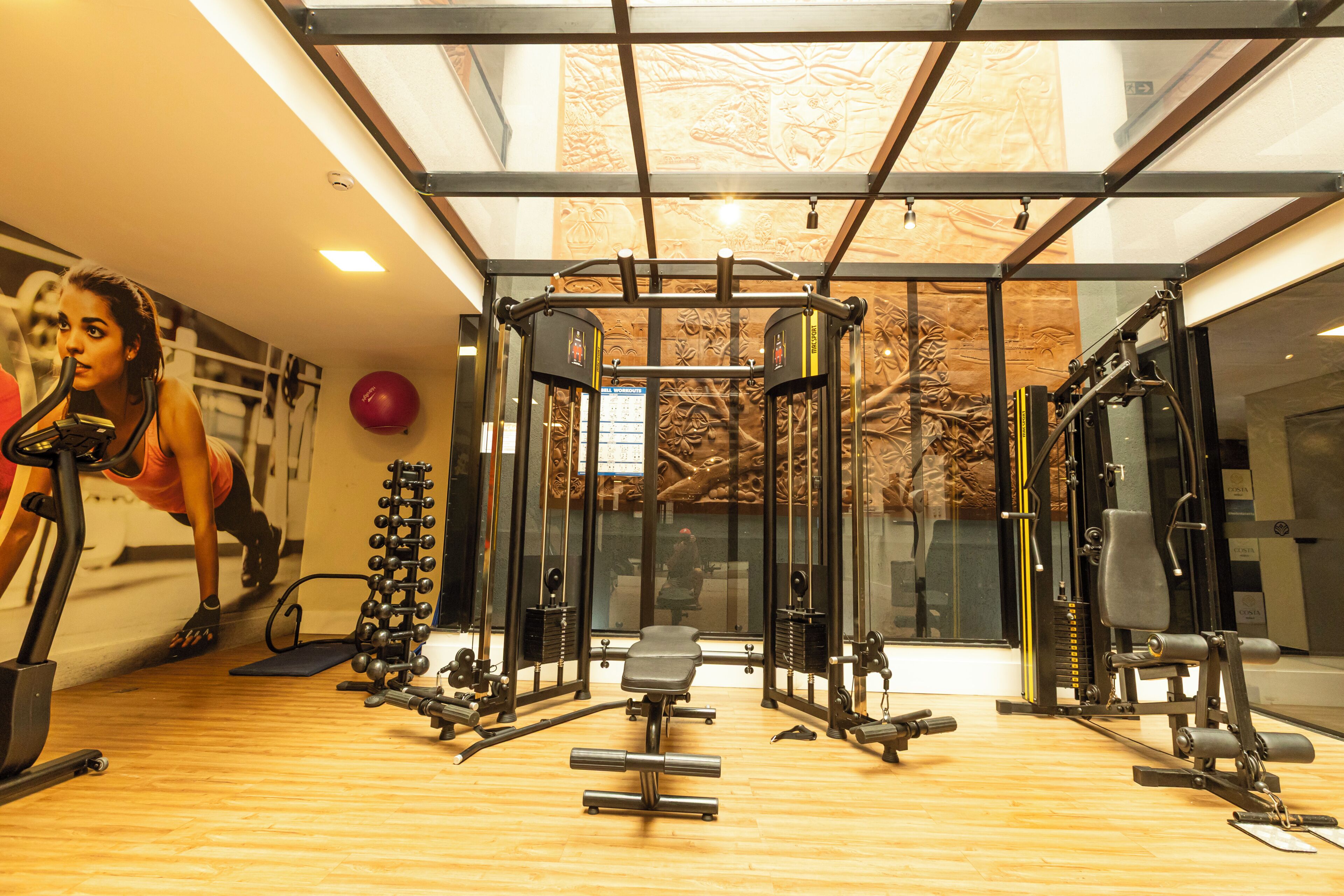 Uma academia com diversos equipamentos de exercício, piso de madeira e uma grande janela com vista para árvores.