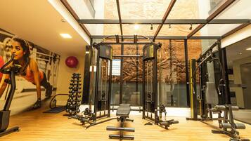 Uma academia com diversos equipamentos de exercício, piso de madeira e uma grande janela com vista para árvores.