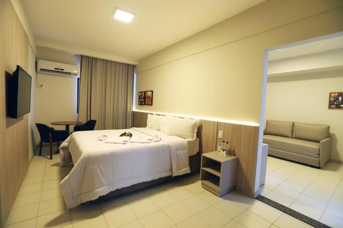 A hotel room with a bed, bedside table, lamp, TV, and a sofa in the adjacent living area.