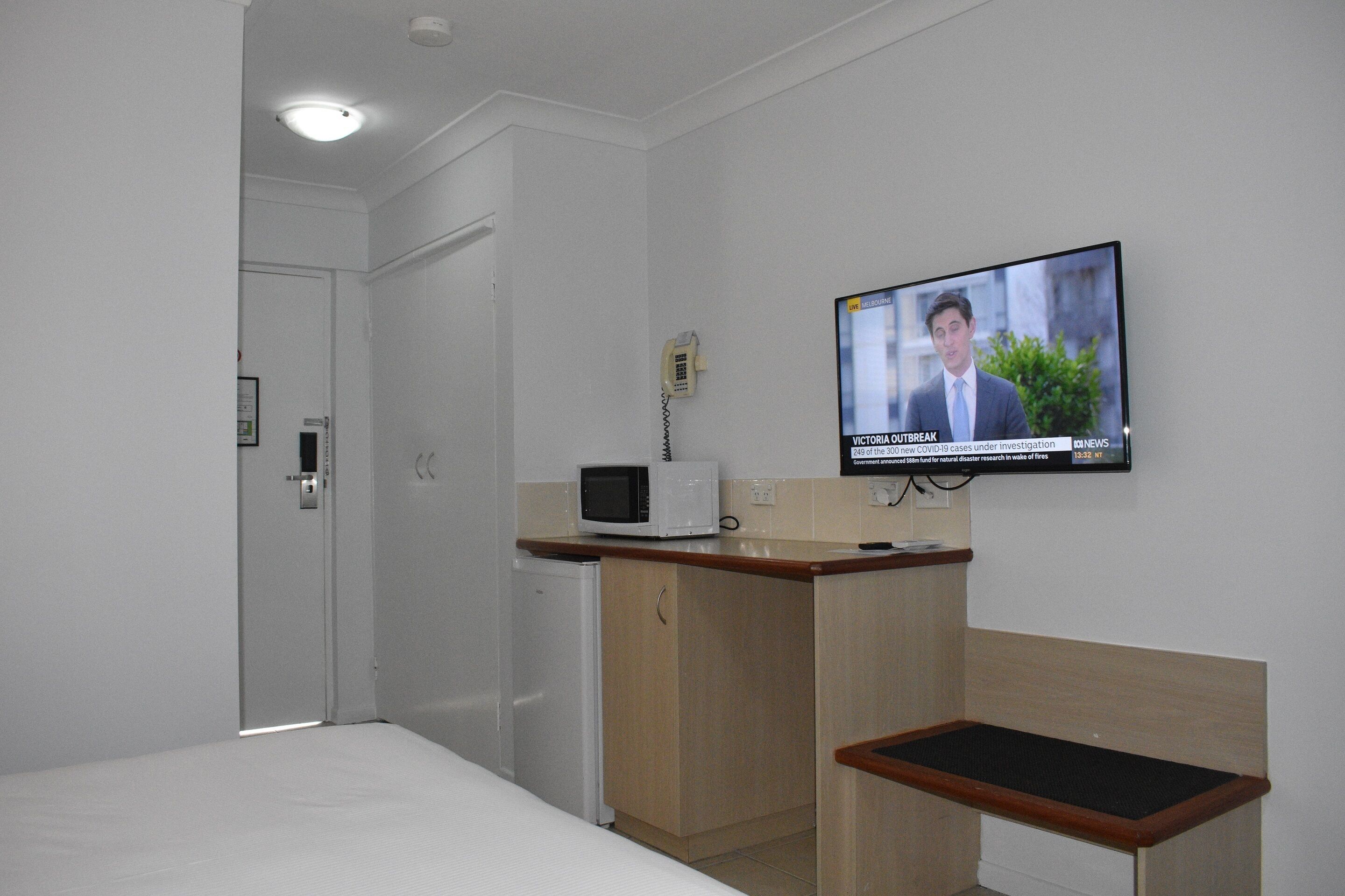 A hotel room with a bed, a small kitchenette with a microwave, a TV displaying a news broadcast, and a door leading to another room.