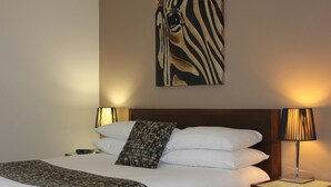 Una cama bien hecha con sábanas blancas y una manta decorativa estampada, una obra de arte con temática de cebra y dos lámparas de noche sobre una mesita de madera.