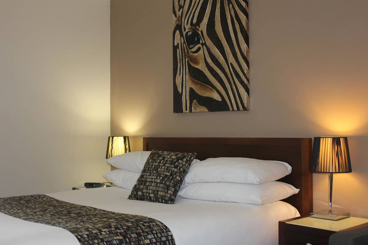A neatly made bed with white linens and a patterned throw blanket, a zebra-themed wall art piece, and two bedside lamps on a wooden nightstand.