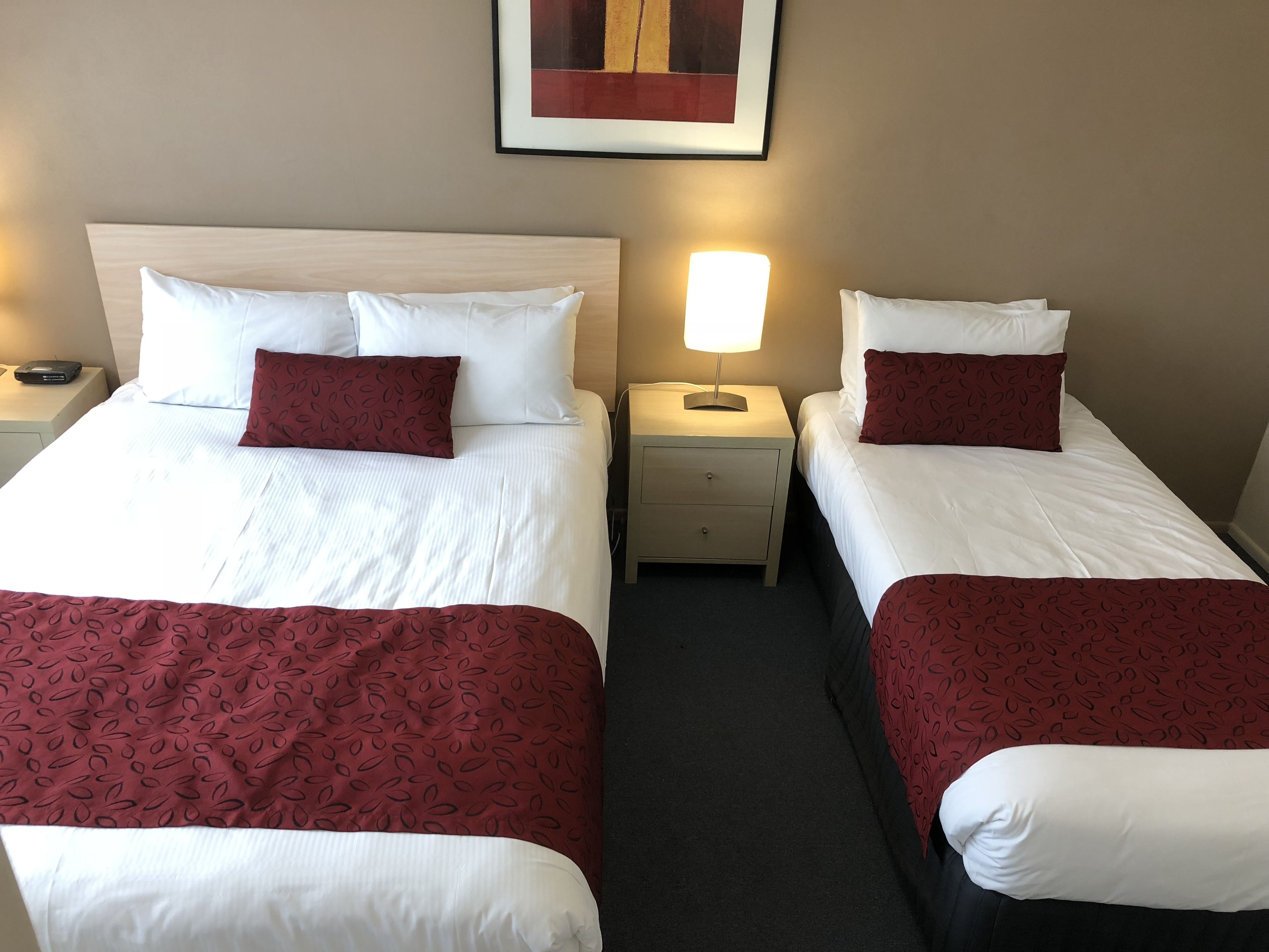 A hotel room with two beds, each with white linens and red patterned bedspreads. There is a nightstand with a lamp between the beds, and a framed picture on the wall above the beds.
