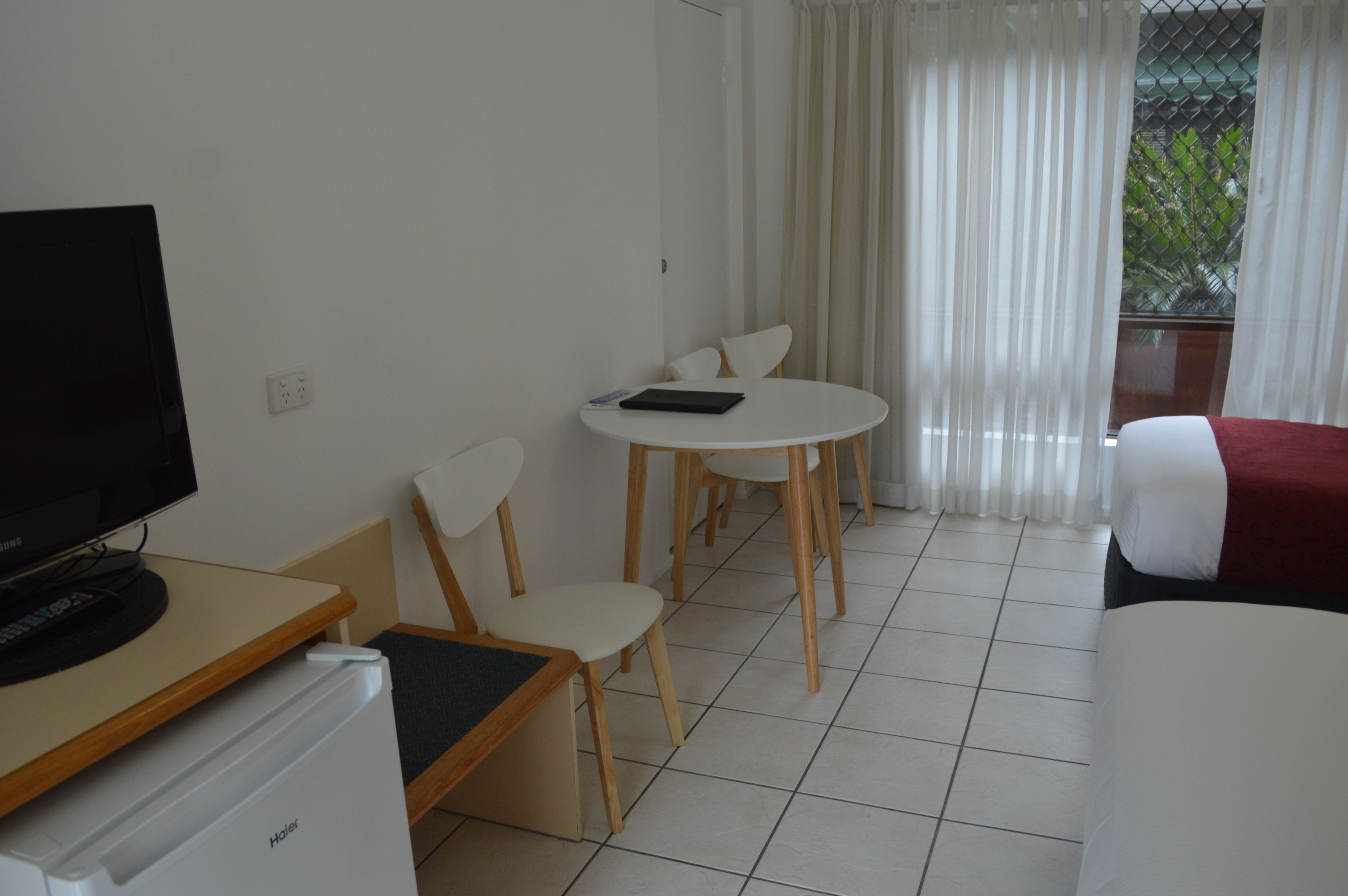 Habitación de hotel con televisión, refrigerador pequeño, mesa de comedor con sillas, cama y ventana con cortinas.