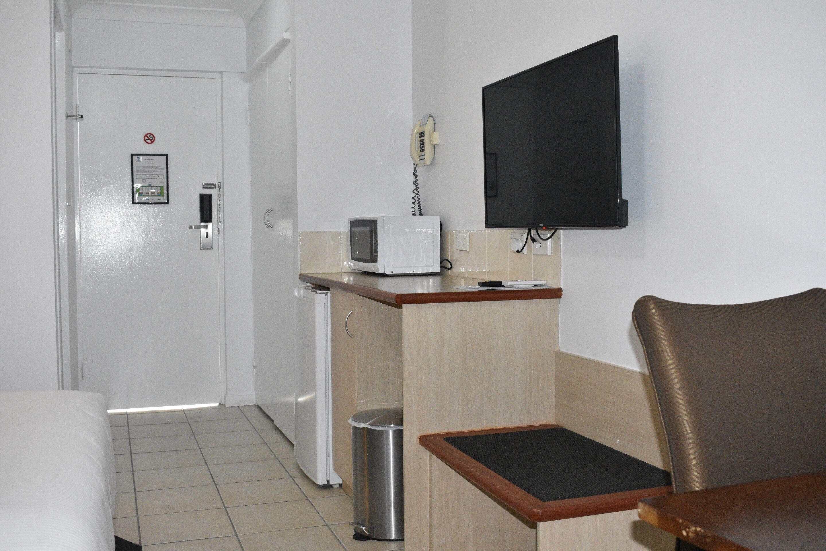A hotel room with a bed, a desk with a computer, a microwave, a trash can, and a TV mounted on the wall.