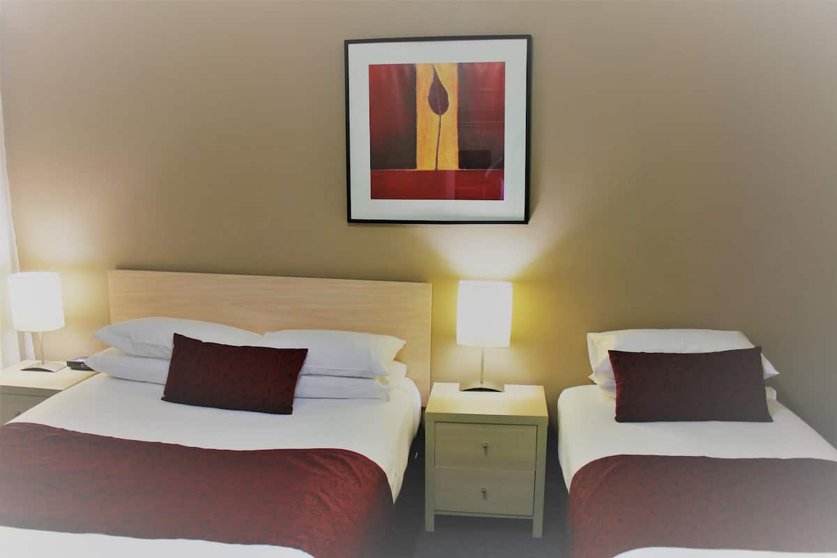 A hotel room with two beds, each with a white duvet and a dark red pillow. There is a framed artwork above the beds, a bedside table with a lamp on each side, and a small drawer unit between the beds.
